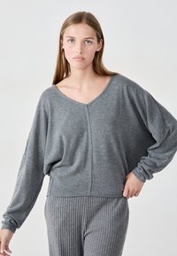 Junge Frau mit langen blonden Haaren, die einen lockeren grauen V-Ausschnitt-Pullover und gerippte graue Hosen trägt, steht vor einem schlichten Hintergrund.