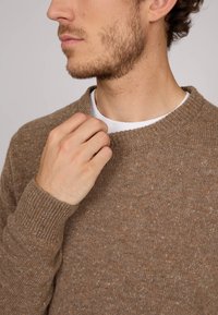 Pull en maille marron avec un col rond, intérieur texturé et des éclats de couleurs claires. Le modèle ajuste la zone du col avec une main visible.