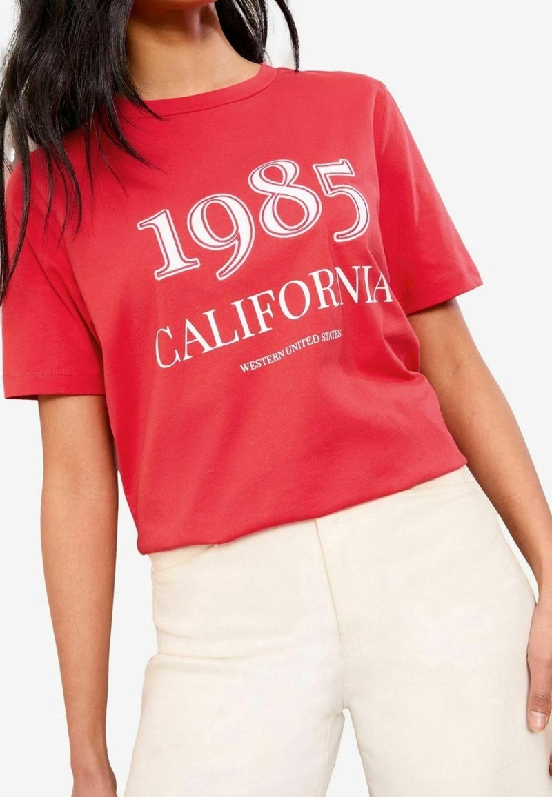 Camiseta de algodón roja con texto blanco que dice "1985 CALIFORNIA WESTERN UNITED STATES." Con mangas cortas y corte relajado. Combinada con pantalones beige.