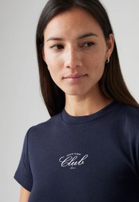 T-shirt azul marinho de algodão com gola circular e mangas curtas, com texto branco "JOIN THE Club" em fonte estilosa na frente.