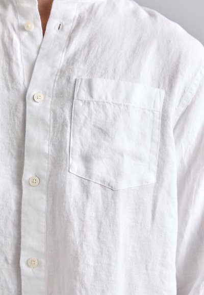 Camicia di lino bianca con pattina abbottonata e tasca. Presenta un tessuto texturizzato, una vestibilità rilassata e fibre naturali morbide. Dettagli minimi.