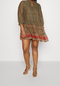 Olijfgroen jurk met driekwart mouwen, bloemenpatroon, kwastje bij de hals en gelaagde zoom met rode accenten. Draagt met gouden hakken.