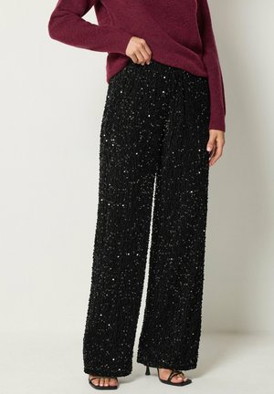 Mujer con pantalones negros de pierna ancha con lentejuelas, camiseta de manga larga burdeos y sandalias negras de tacón abierto.