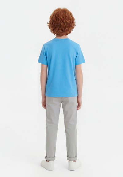 Copil cu păr roșcat și creț, purtând un tricou albastru, pantaloni gri deschis suflecați și adidași albi, stând cu spatele spre privitor pe un fundal alb.
