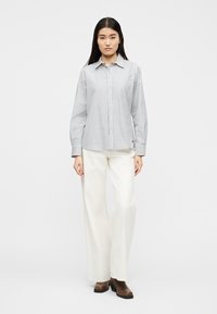 Γυναίκα που φοράει λευκό πουκάμισο με μακριά μανίκια και λεπτές μαύρες κάθετες ρίγες, λευκό παντελόνι φαρδύ στη μπότα και καφέ μποτάκια αστραγάλου.