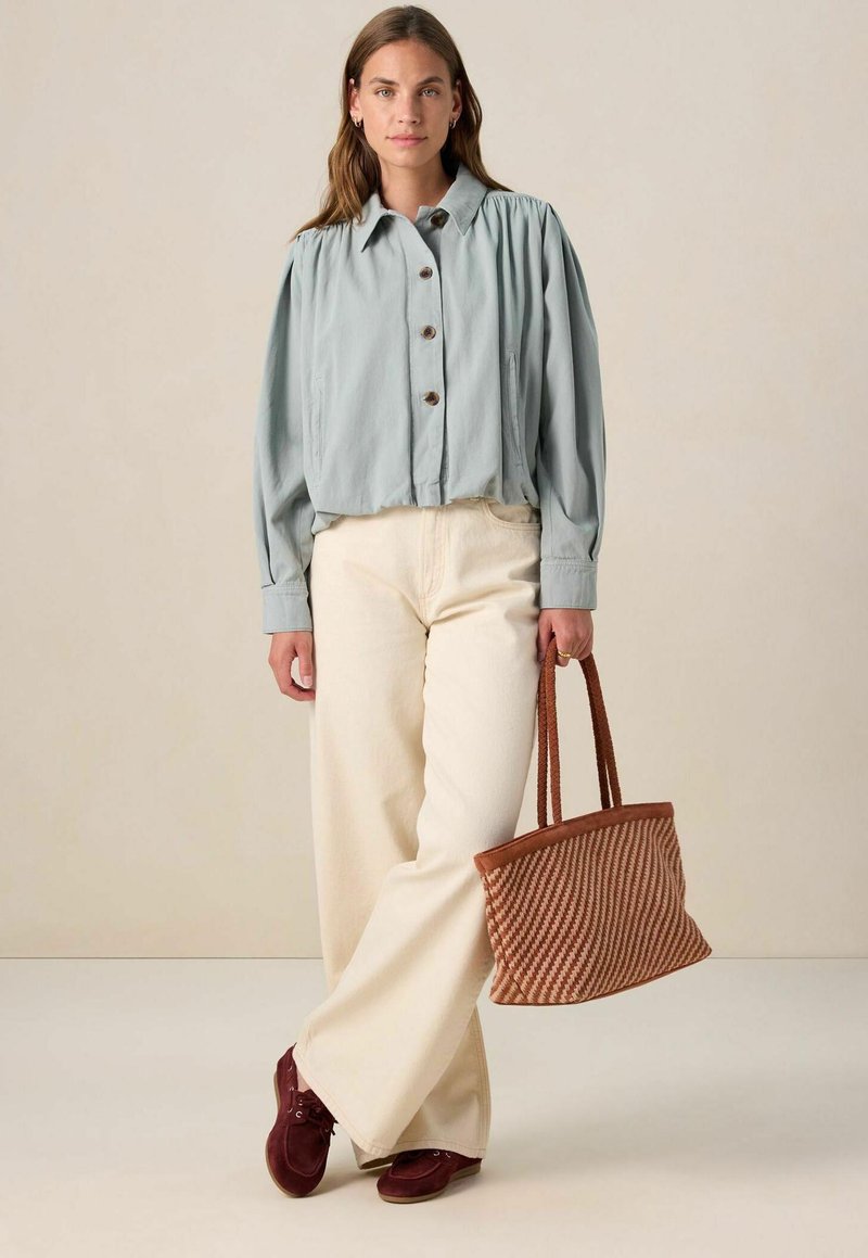 Veste bleue à boutons avec des manches bouffantes, pantalon large crème, et un sac en osier marron. Des chaussures bordeaux complètent la tenue.