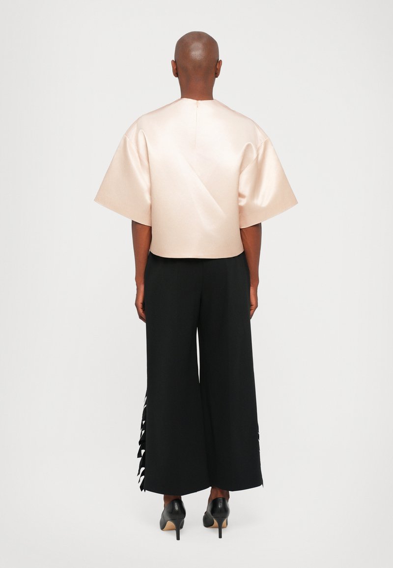 Helles rosa Satinbluse mit weiten Ärmeln, kombiniert mit schwarzen, weit geschnittenen Hosen mit seitlichen Rüschen. Schwarze High Heels runden den Look ab.