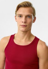 Rode geribde tanktop met brede bandjes, voorzien van een aansluitend ontwerp en een soepele textuur. Model toont de top tegen een neutrale achtergrond.