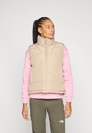 Donna con capelli intrecciati che indossa un gilet imbottito beige sopra una felpa rosa e pantaloni verde oliva, guarda verso destra su uno sfondo neutro.
