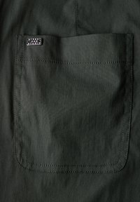 Tessuto verde scuro con una texture liscia, caratterizzato da una tasca a toppa e un'etichetta nera con la scritta "STREET ONE STUDIO" cucita sul tessuto.