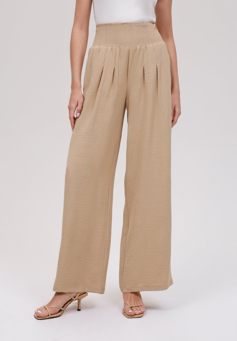 Beige wide-leg pants with a textured finish, featuring a high smocked waist and pleats for added volume. Paired with strappy heels.