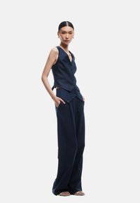 Marineblauwe getailleerde vest met knopen en een laag uitgesneden halslijn, gecombineerd met wijde broekspijpen. Strak, soepel materiaal met een minimalistisch ontwerp.