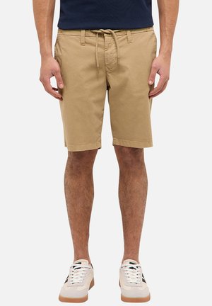 Mężczyzna ubrany w beżowe szorty ze ściągaczem, białe trampki z gumową podeszwą i granatową koszulę, stojący na tle jednolitego jasnego tła.