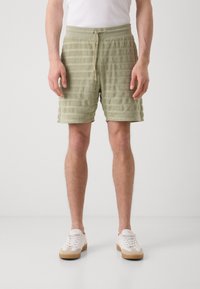 Groene gestreepte handdoeken shorts met een elastische tailleband en trekkoord. De textuur is zacht, met horizontale lijnen voor het design. Gekleed met witte sneakers.