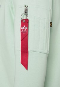 Felpa verde chiaro con tasca con zip, caratterizzata da un'etichetta rossa con testo e logo bianchi. Texture liscia e dettagli di cucitura in evidenza.