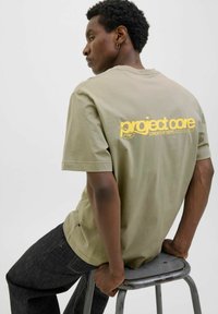 Olivengrøn, kortærmet t-shirt i blødt stof, med en stor gul "project core" grafik på ryggen. Båret med mørke denimbukser.