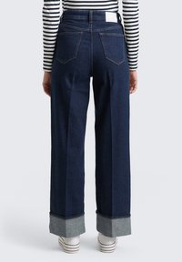Szerokie, ciemnoniebieskie jeansy z denimu z podwiniętymi mankietami, pięcioma kieszeniami oraz czystym tyłem z białą metką przy pasku.
