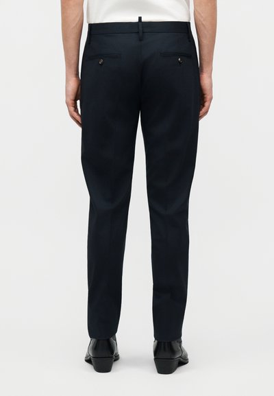 Άνδρας που φοράει μαύρο ραμμένο παντελόνι, μαύρα παπούτσια και λευκό πουκάμισο, στέκεται με τα χέρια χαλαρά στα πλευρά, οπισθοσκοπική όψη.