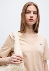 Νεαρή γυναίκα που φοράει μπεζ T-shirt με λαιμόκοψη V και πράσινο λογότυπο κροκόδειλου, κρατώντας ιμάντα τσάντας με λευκή αφράτη υφή πάνω στον ώμο της, σε λευκό φόντο.