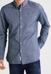 Blå skjorta med knappar i vävt tyg med en texturerad yta. Har krage, långa ärmar och en liten logotyp på bröstet.