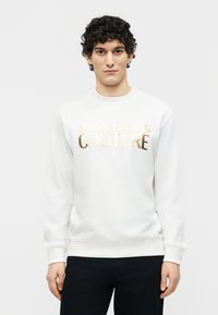 Νεαρός άνδρας με σγουρά σκούρα μαλλιά που φοράει λευκό φούτερ με χρυσά γράμματα "Versace Jeans Couture", στεκόμενος μπροστά από ένα απλό φόντο.