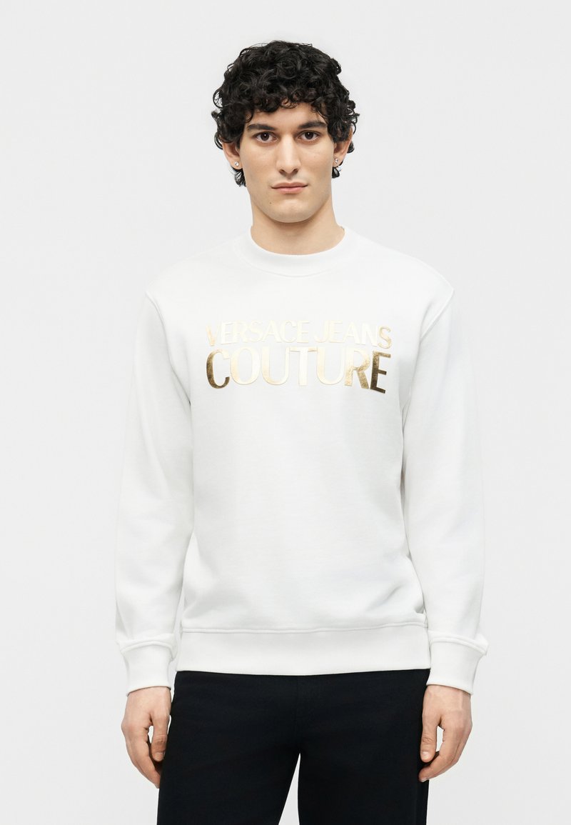 Νεαρός άνδρας με σγουρά σκούρα μαλλιά που φοράει λευκό φούτερ με χρυσά γράμματα "Versace Jeans Couture", στεκόμενος μπροστά από ένα απλό φόντο.