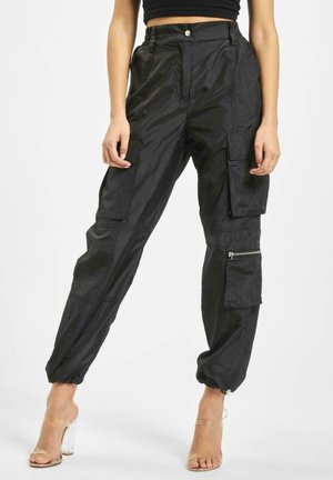 Femme porte un pantalon cargo noir taille haute avec plusieurs poches et des poignets élastiqués, assorti à des sandales à talons transparents.
