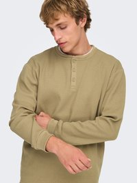 Chemise kaki à manches longues en tricot gaufré avec un col rond et une patte de boutonnage à trois boutons ; elle présente des poignets côtelés et un tissu texturé.