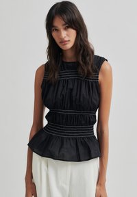 Juodas peplumo topas su struktūrizuotu priekiniu sijonu, pasižymintis sukaupta medžiaga ir subtiliomis baltomis siūlėmis krūtinėje ir liemenyje.