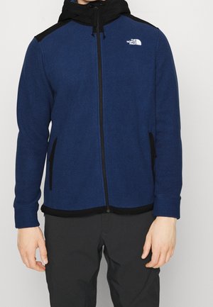 Mann trägt eine blau-schwarze Fleecejacke mit Reißverschluss, Kapuze und The North Face-Logo auf der linken Brust.