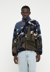 Jachetă din fleece cu guler înalt, având un model de peisaj montan în culorile albastru, verde și maro, și un fermoar negru.