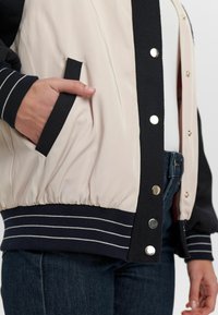 Veste beige clair avec une manche bleu marine et un bord-côte au poignet. Comprend des boutons-pression, des poches latérales et des rayures blanches à l'ourlet.