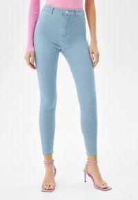 Jeans ajustados de cintura alta azul claro, confeccionados en denim, con cierre frontal de botón y textura suave. Combinados con sandalias de tacón alto moradas.