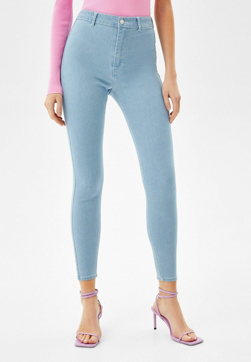 Jeans ajustados de cintura alta azul claro, confeccionados en denim, con cierre frontal de botón y textura suave. Combinados con sandalias de tacón alto moradas.