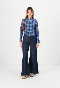 Kvinne som står iført blå rutete skjorte, brede mørke jeans, beige sneakers og brun skulderveske foran en plain hvit bakgrunn.