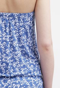 Top floreale blu con scollatura elastica arricciata, caratterizzato da motivi di fiori bianchi su un tessuto morbido e leggero. La vista posteriore mostra il design.