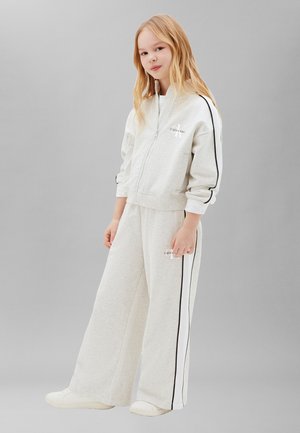 Noor tüdruk seisab heledas hallis Calvin Kleini dressis mustade triibudega ja valgete tossudega ühevärvilise halli taustaga.