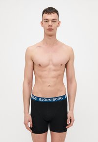 Black boxer briefs with a blue waistband featuring "BJÖRN BORG" logo. Smooth fabric, form-fitting design, mid-thigh length.