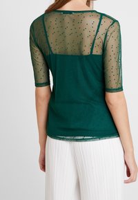 Top verde de malla con mangas cortas, que presenta pequeños patrones de puntos. Tejido semitransparente y diseño de espalda sólida combinado con pantalones blancos con pliegues.