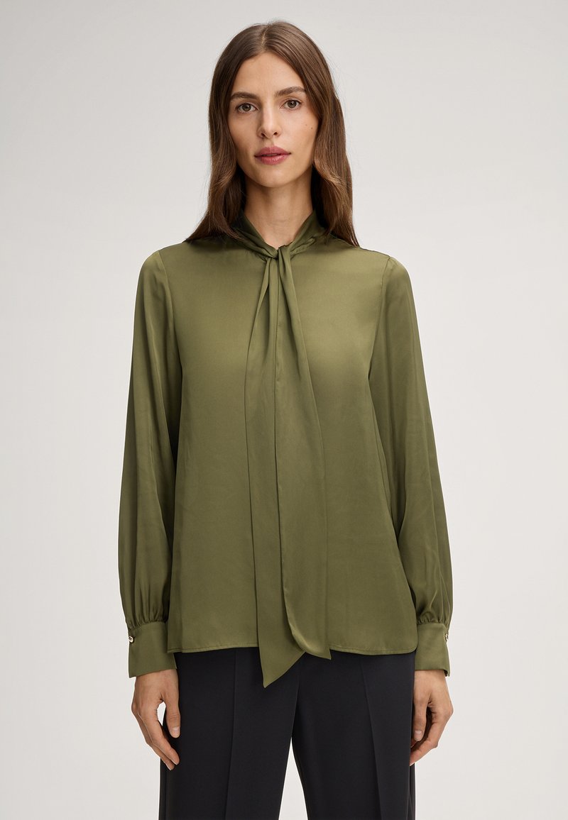 Blusa di seta verde oliva con scollatura a fiocco, maniche lunghe e polsini abbottonati. Tessuto liscio e vestibilità drappeggiata accentuata da un leggero luccichio.