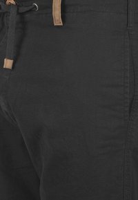 Pantalones de tela negra con textura suave, con cierre de cordón y detalles en cuero marrón claro en la cintura.