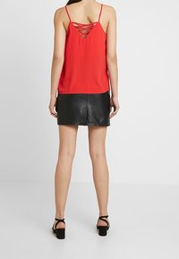 Červený bezramenný top s kříženým designem na zádech, doplněný černou koženou mini sukní. Modelka má na sobě černé sandály na blokovém podpatku.