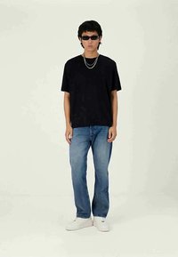 Νεαρός που φορά μαύρο t-shirt, μπλε τζιν, λευκά αθλητικά παπούτσια, σκούρα γυαλιά ηλίου και ασημένια αλυσίδα στο λαιμό, ποζάροντας σε απλό φωτεινό φόντο.