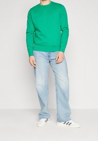 Felpa verde girocollo in cotone, abbinata a jeans chiari a taglio morbido e sneakers bianche con strisce nere. Il modello si trova davanti a uno sfondo neutro.