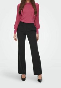 Haut rose à manches longues avec poignets côtelés, assorti à un pantalon noir sur mesure. Talons noirs pointus complètent la tenue. Textures lisses et ajustées.