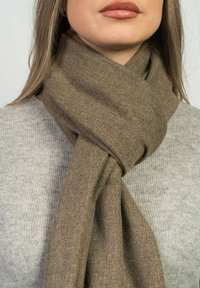 En brun ullsjal snurrat runt halsen, med en mjuk, texturerad yta. Bärs över en ljusgrå stickad tröja.