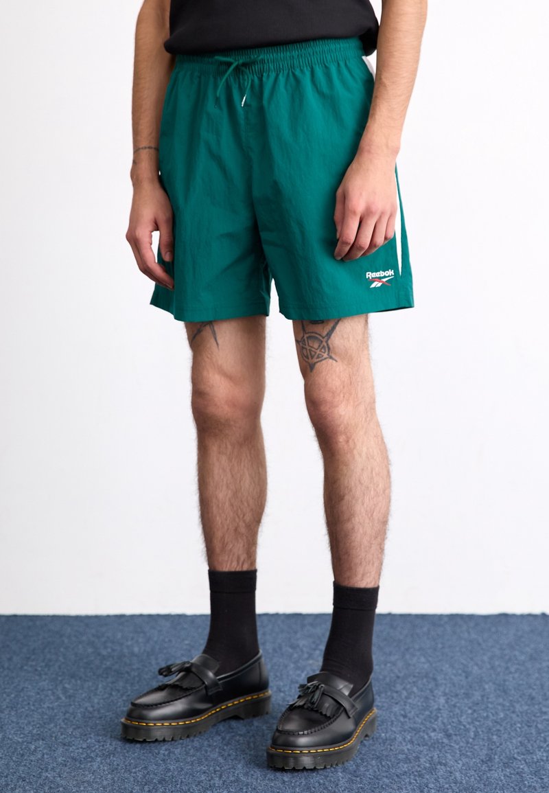 Tyrkysové plavky s elastickým pasem a šňůrkou. Obsahují logo Reebok; nošeny s černými ponožkami a platformovými mokasíny.