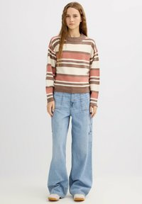 Jersey de rayas en beige, rosa y marrón. Vaqueros cargo de pierna ancha en azul claro con bolsillos laterales. Modelo de pie sobre un fondo claro.