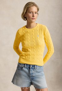 Γυναίκα που φοράει κίτρινο πουλόβερ με πλέξη καλώδιο και ανοιχτόχρωμα σορτς denim, στέκεται με τα χέρια πίσω από την πλάτη της μπροστά σε ουδέτερο φόντο.