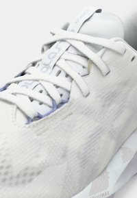 Λευκό αθλητικό παπούτσι με υφή στην πάνω μέρος, λεπτούς κορδόνια και ανοιχτό μωβ λεπτομέρειες. Διαθέτει καμπύλη σόλα με σχεδιαστικό μοτίβο από κάτω.